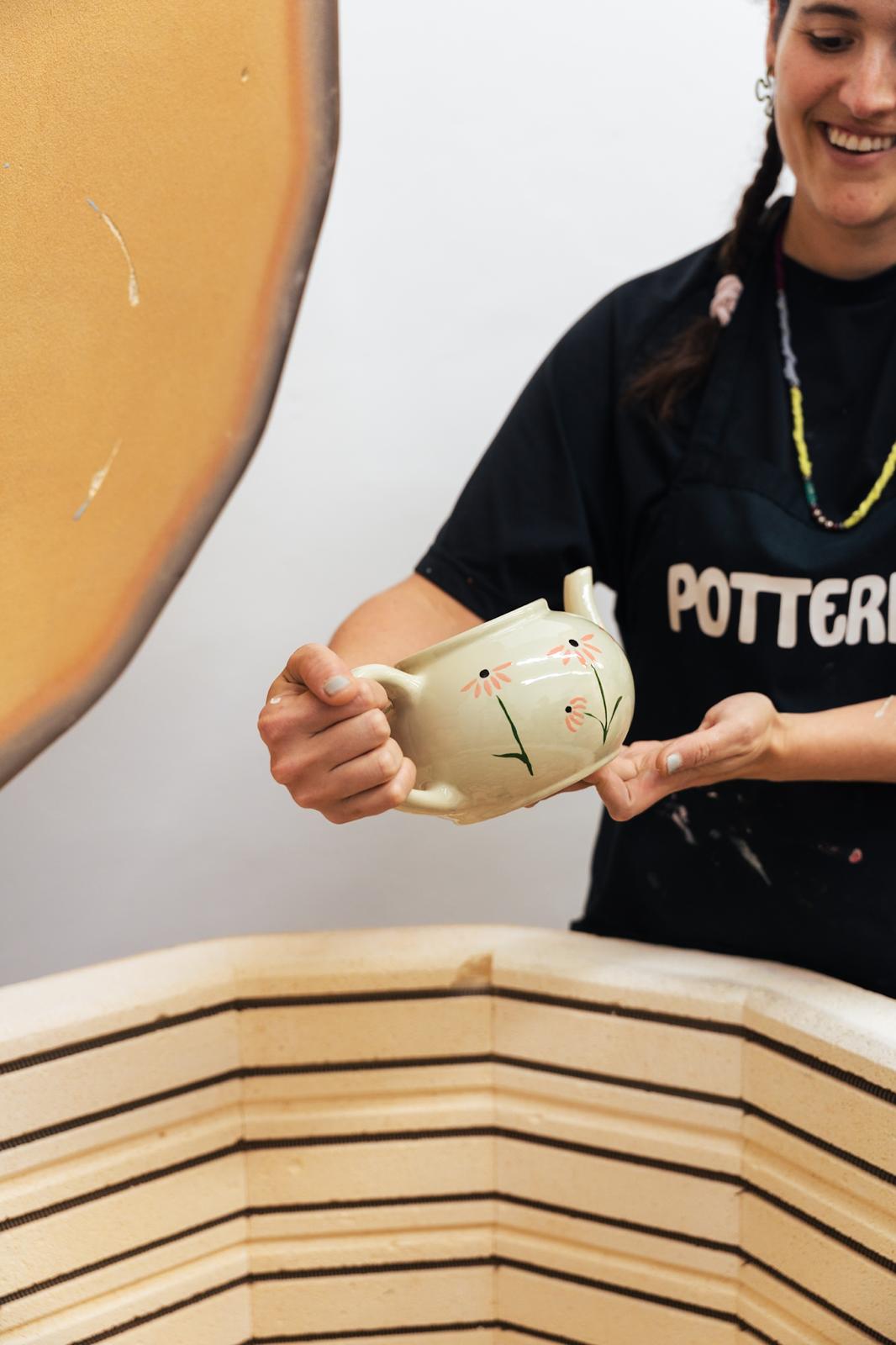 Jenna holding a floral-painted teapot beside the PotterMe kiln