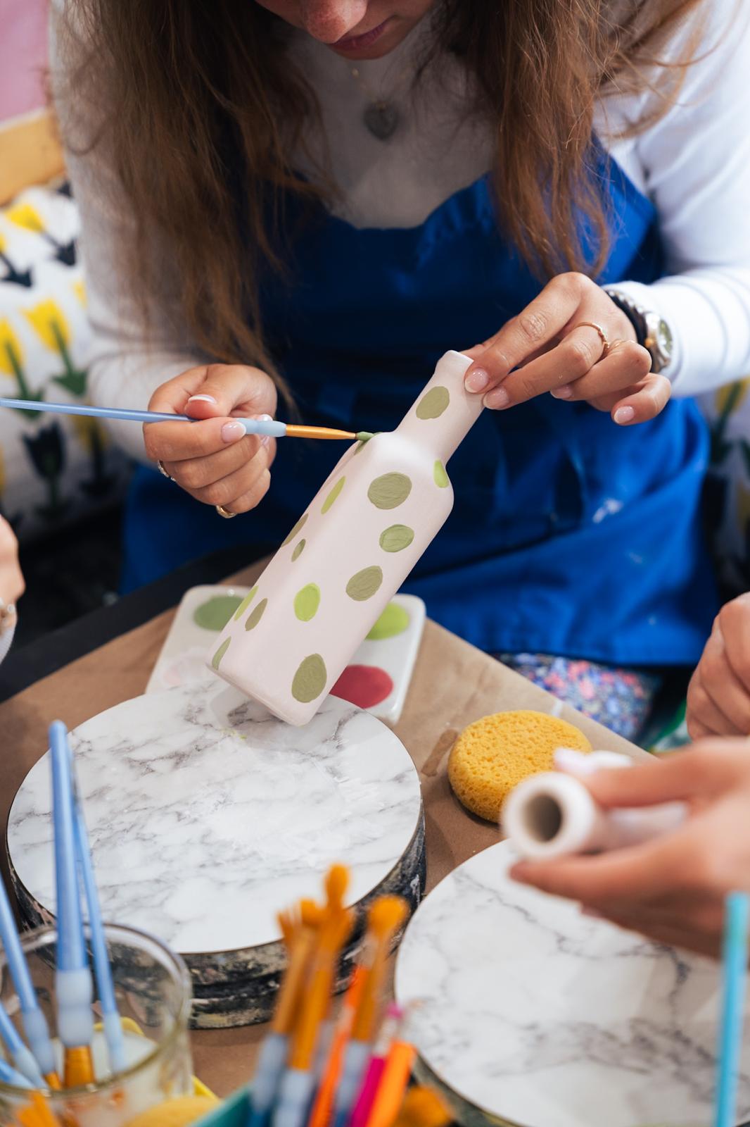 A guest painting green dots on a ceramic bottle at a shared PotterMe table